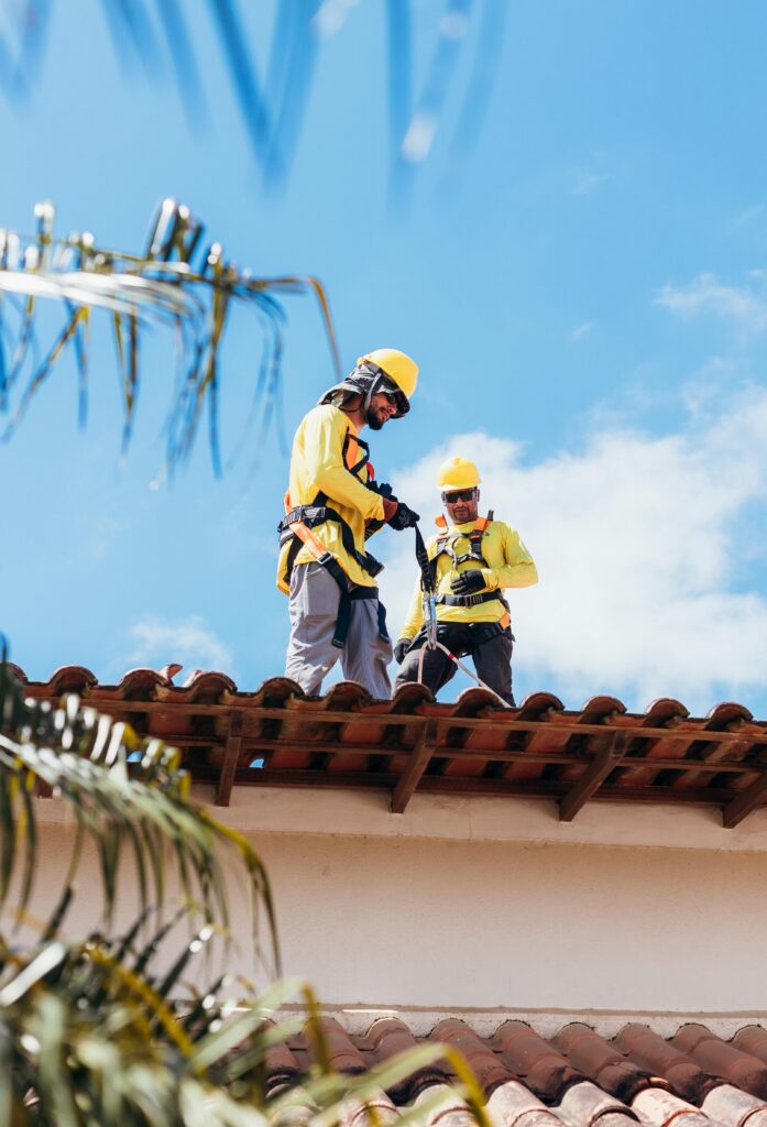 Two professional roofers wearing safety harnesses and helmets working together on a residential roof.